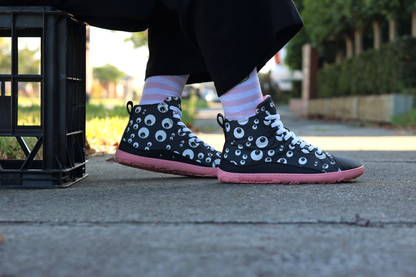 PaperKrane barefoot eyeball boots on legs with stripey socks, on a footpath. The person is sitting on a crate wearing boots that are black with cartoon eyeballs, elastic laces, they're a foot-shaped, zero-drop design, on a salmon coloured sole. 