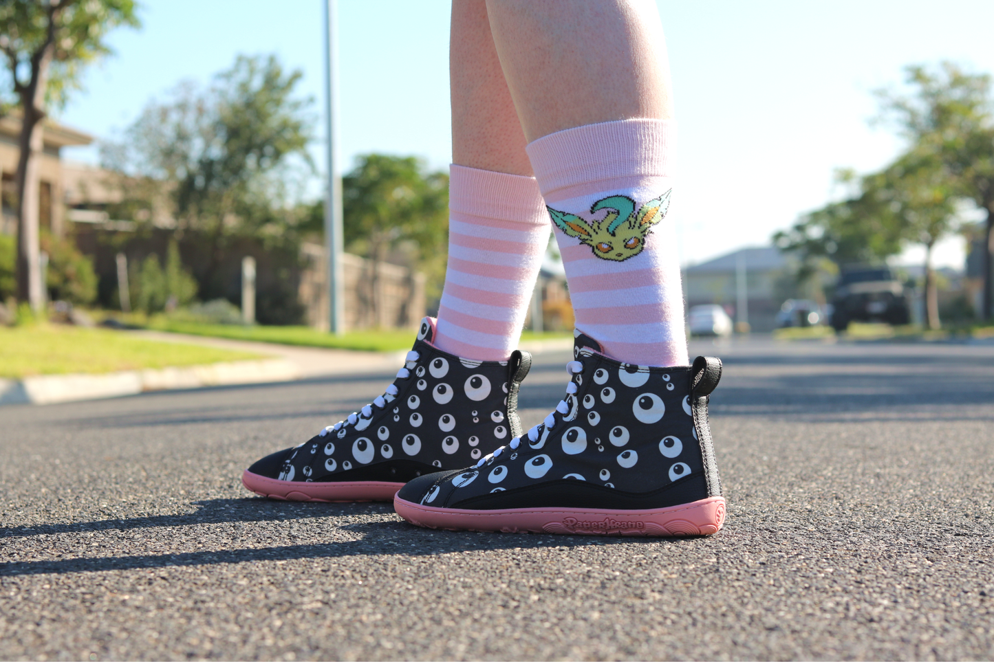 PaperKrane barefoot eyeball boots on legs with stripey socks, on a street facing to the left. The boots are black with cartoon eyeballs, elastic laces, they're a foot-shaped, zero-drop design, on a salmon coloured sole. 