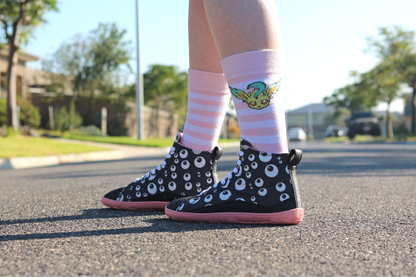 PaperKrane barefoot eyeball boots on legs with stripey socks, on a street facing to the left. The boots are black with cartoon eyeballs, elastic laces, they're a foot-shaped, zero-drop design, on a salmon coloured sole. 