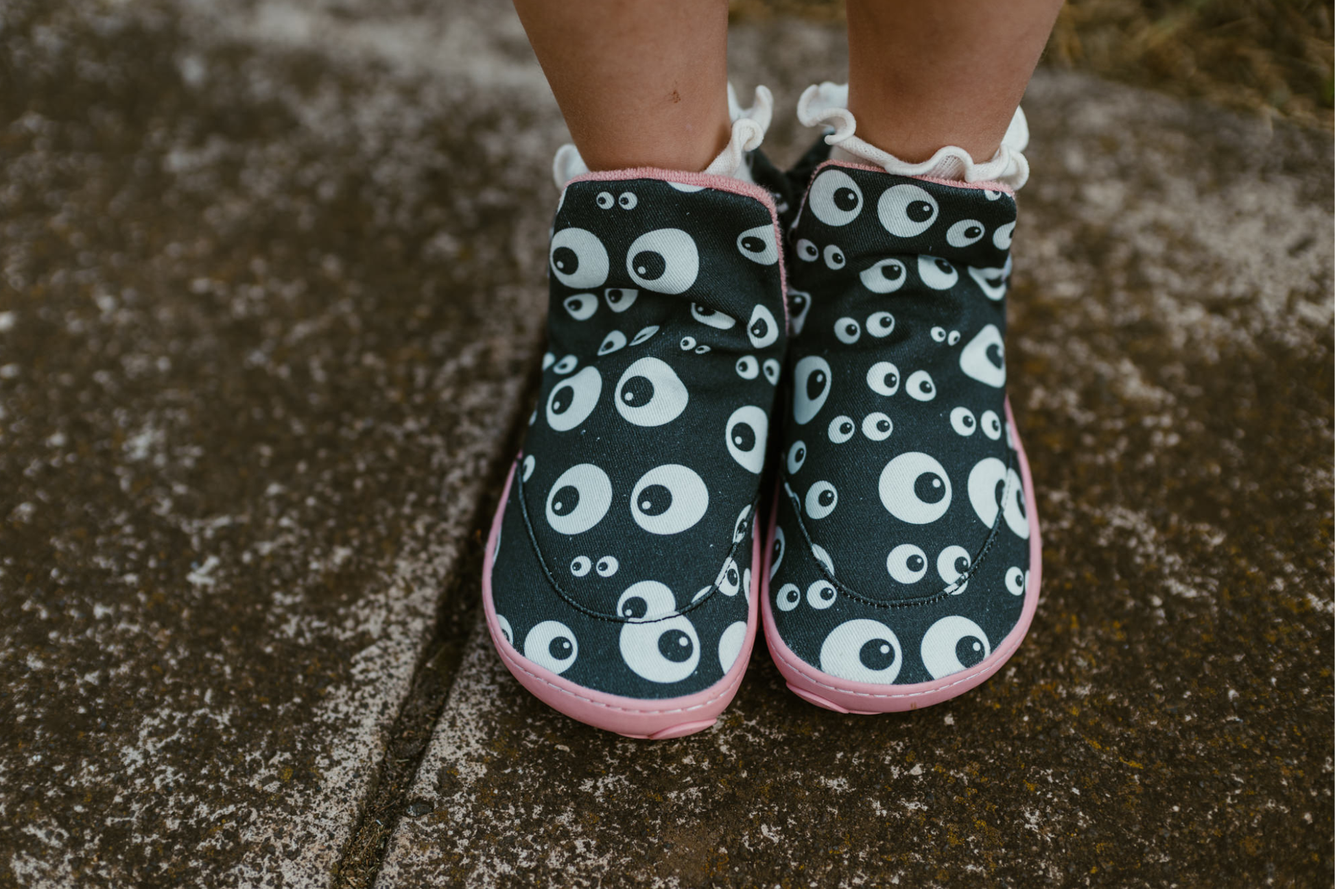 Above view of PaperKrane barefoot eyeball signature style high tops on little legs with white socks, standing on a footpath. The slip on shoes are black with cartoon eyeballs, elastic ankles, they're a foot-shaped, zero-drop design, on a salmon coloured sole. 