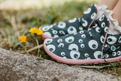 Close up side view of PaperKrane barefoot eyeball signature style high tops on little legs with white socks, sitting next to a footpath. The slip on shoes are black with cartoon eyeballs, elastic ankles, they're a foot-shaped, zero-drop design, on a salmon coloured sole. 