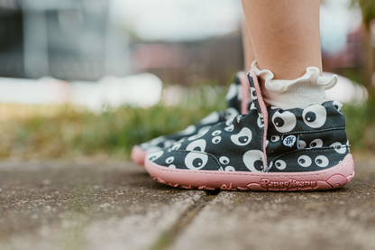 PaperKrane barefoot eyeball signature style high tops on little legs with white socks, standing on a footpath. The slip on shoes are black with cartoon eyeballs, elastic ankles, they're a foot-shaped, zero-drop design, on a salmon coloured sole. 