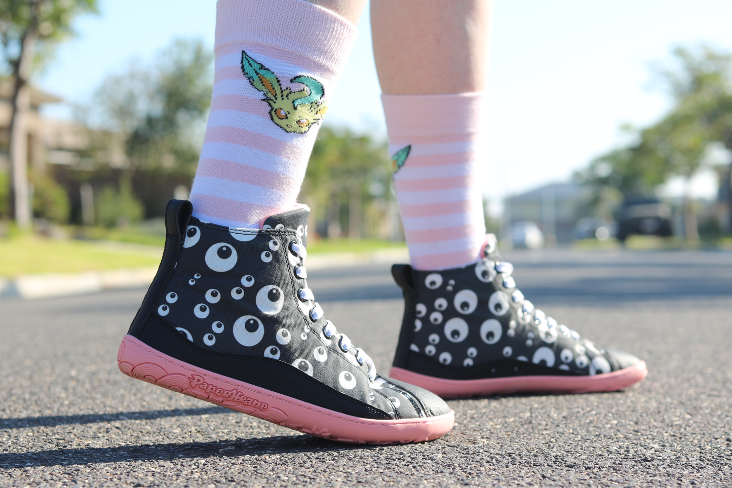 PaperKrane barefoot eyeball boots on legs with stripey socks, on a street. The boots are black with cartoon eyeballs, elastic laces, they're a foot-shaped, zero-drop design, on a salmon coloured sole. 