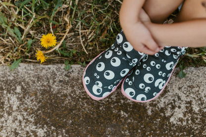 Abvoe view of PaperKrane barefoot eyeball signature style high tops on little legs, sitting on grass next to a footpath. The slip on shoes are black with cartoon eyeballs, elastic ankles, they're a foot-shaped, zero-drop design, on a salmon coloured sole. 