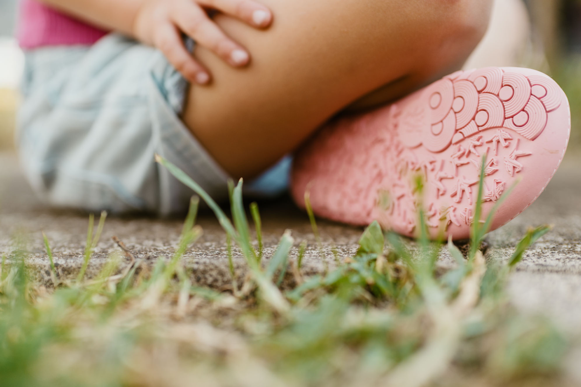 A little peep at the sole of a pair of PaperKrane barefoot eyeball signature style high tops on little legs, sitting on a footpath. The slip on shoes are black with cartoon eyeballs, elastic ankles, they're a foot-shaped, zero-drop design, on a salmon coloured sole - which we can clearly see in this picture. 