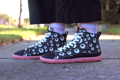 PaperKrane barefoot eyeball boots on legs with stripey socks and black pants, on a footpath. The boots are black with cartoon eyeballs, elastic laces, they're a foot-shaped, zero-drop design, on a salmon coloured sole. 