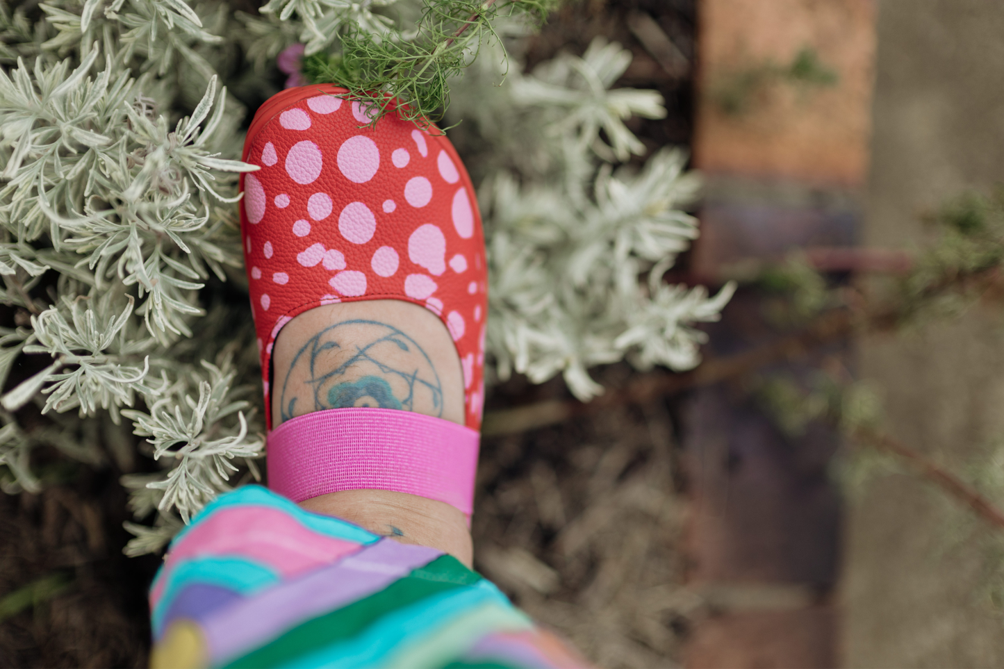 Person wearing one PaperKrane red leather foot-shaped mary jane shoe with pink spots and shrubbery in the background. 