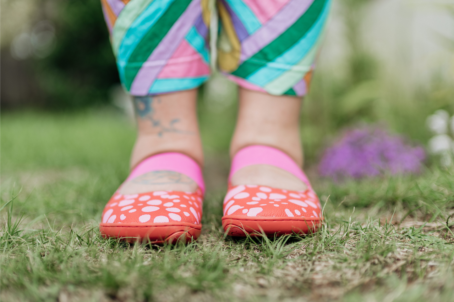 Person wearing colourful pants, and red leather PaperKrane mary jane shoes with pink spots painted on, and pink elastic straps, on grass. 