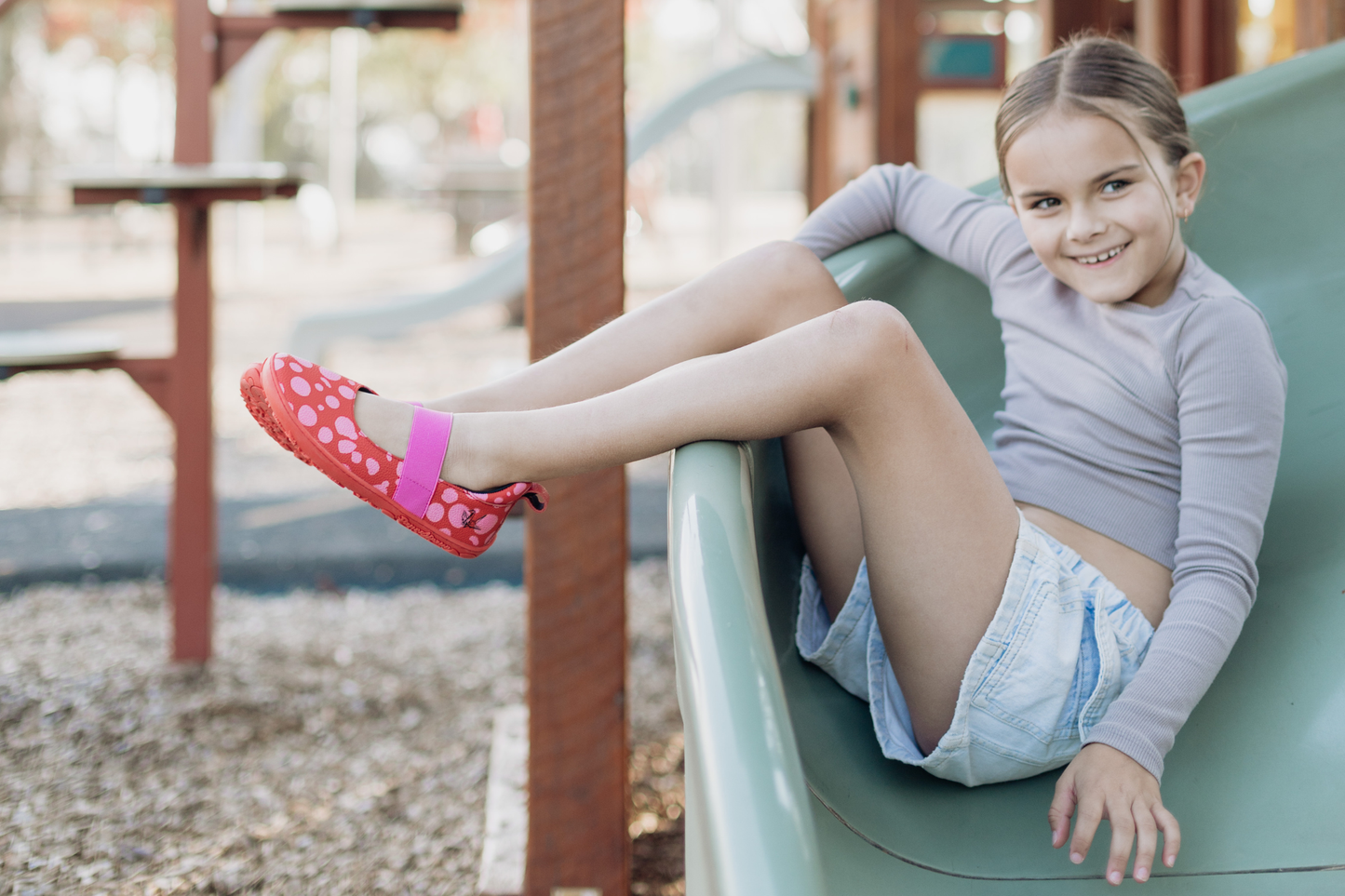 Child on a slide wearing red leather PaperKrane barefoot mary jane shoes with pink dots and elastic strap and red rubber soles.