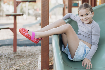Child on a slide wearing red leather PaperKrane barefoot mary jane shoes with pink dots and elastic strap and red rubber soles.
