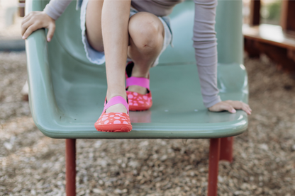 Child's feet wearing wide toebox red leather PaperKrane mary jane shoes with pink dots and elastic straps on a slide.