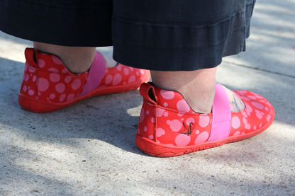 Back view of Red leather foot-shaped mary jane shoes with painted bubblegum pink spots and pink elastic strap on high volume adult feet.