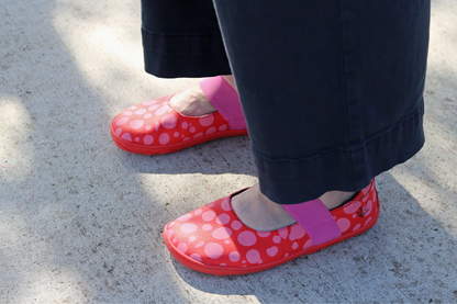 Front angled view of Red leather foot-shaped mary jane shoes with painted bubblegum pink spots and pink elastic strap on high volume adult feet.