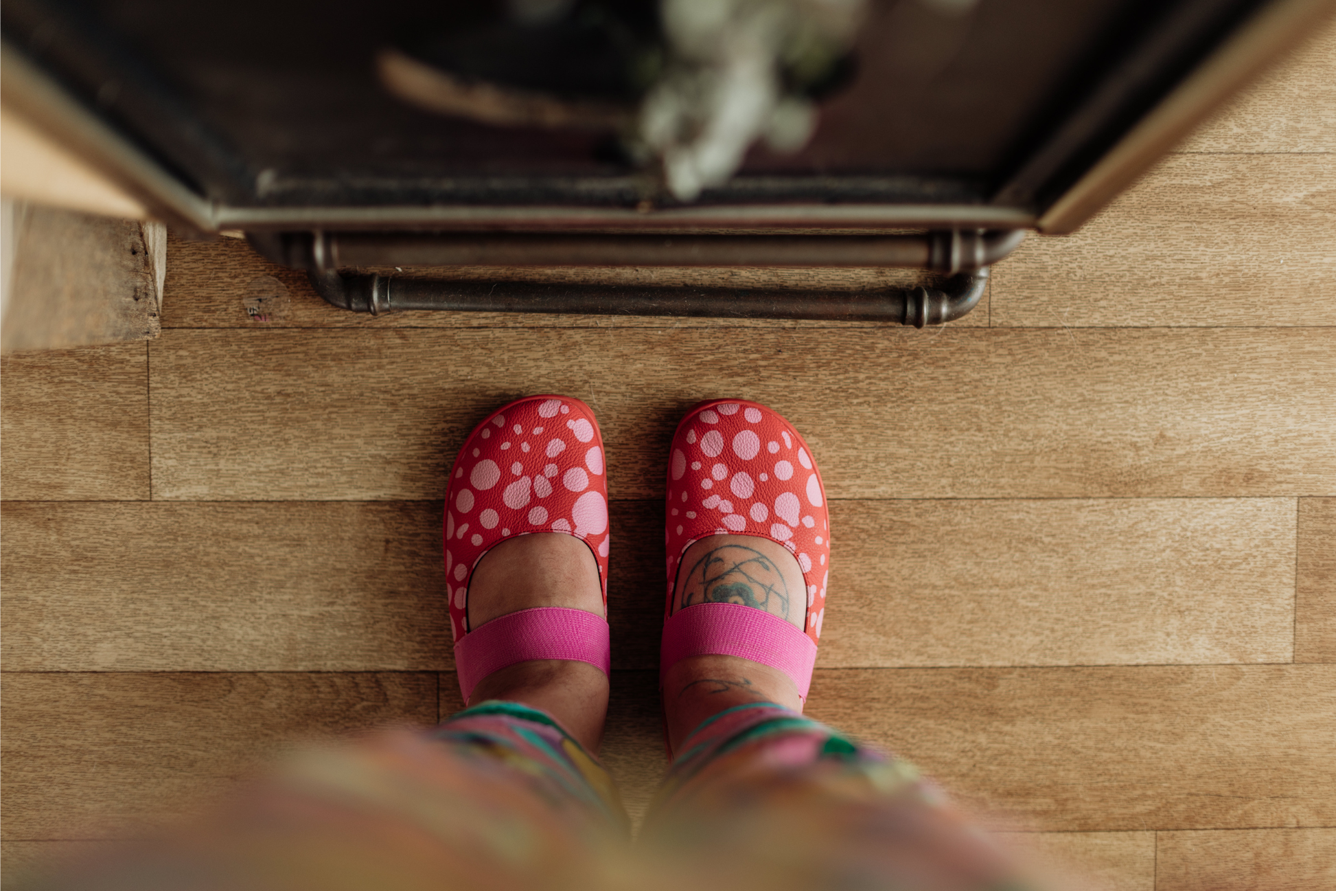 Above view of Red leather foot-shaped mary jane shoes with painted bubblegum pink spots and pink elastic strap on high volume adult feet. They're on a wooden floor. 