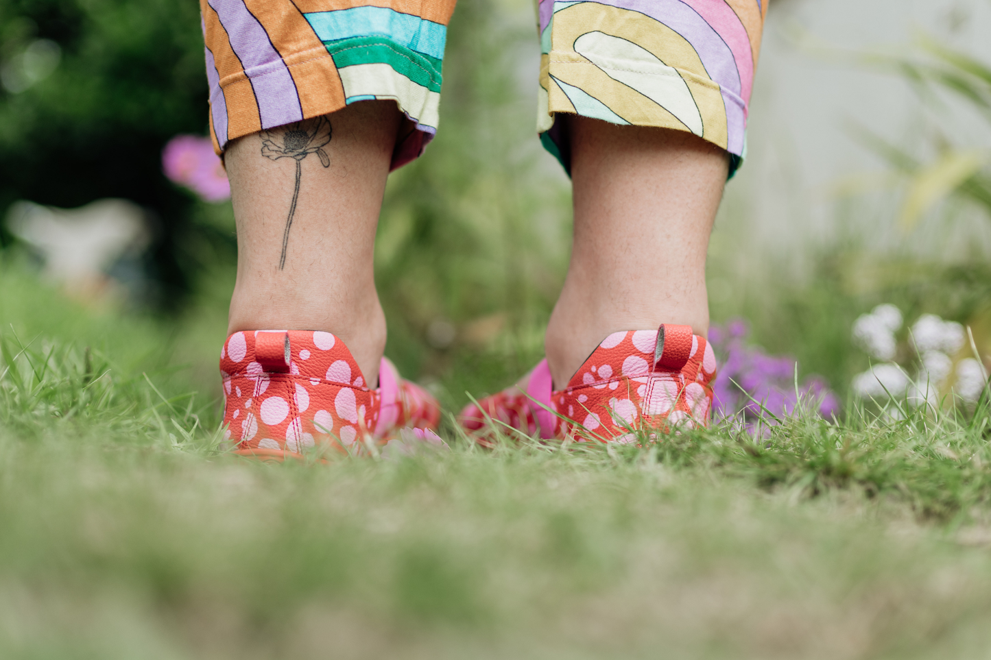 Person wearing colourful pants and red leather PaperKrane mary jane shoes with pink spots standing on grass.
