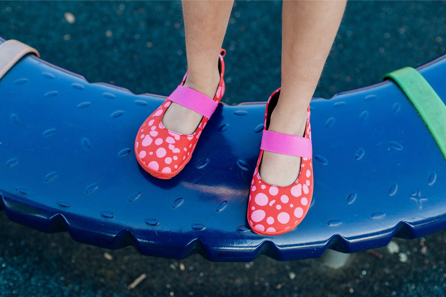 Child's feet wearing foot-shaped red leather PaperKrane mary jane shoes with pink dots and elastic straps on a blue playground surface.