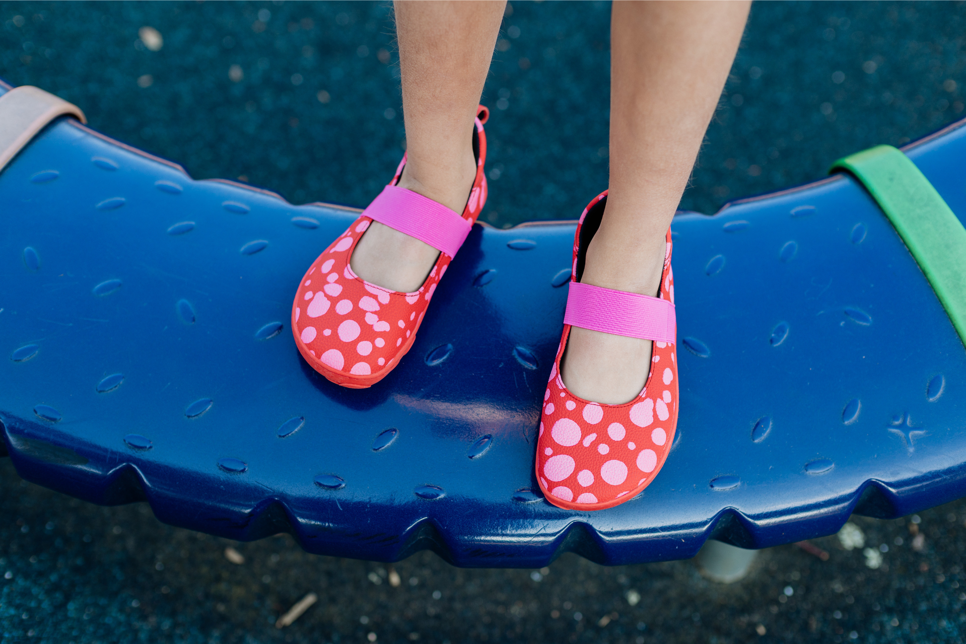 Child's feet wearing foot-shaped red leather PaperKrane mary jane shoes with pink dots and elastic straps on a blue playground surface.