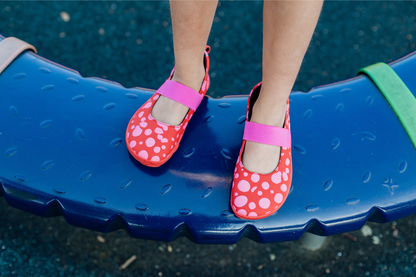 Child's feet wearing foot-shaped red leather PaperKrane mary jane shoes with pink dots and elastic straps on a blue playground surface.