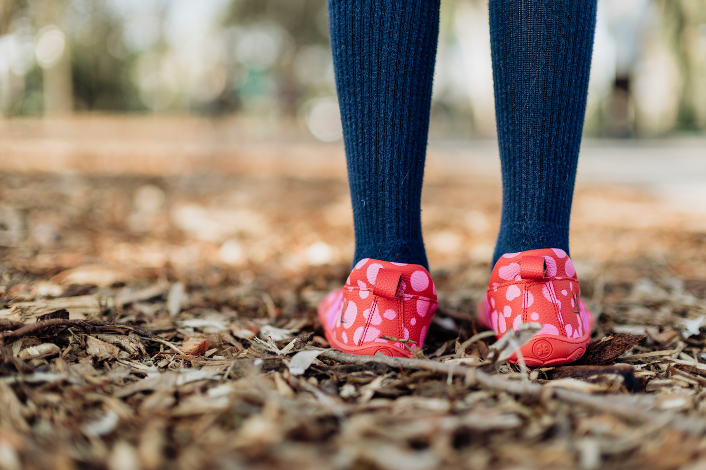 Child's feet in ling blue socks wearing barefoot red leather PaperKrane mary jane shoes with pink dots on bark.