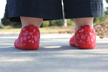 Back view of Red leather foot-shaped mary jane shoes with painted bubblegum pink spots and pink elastic strap on high volume adult feet.