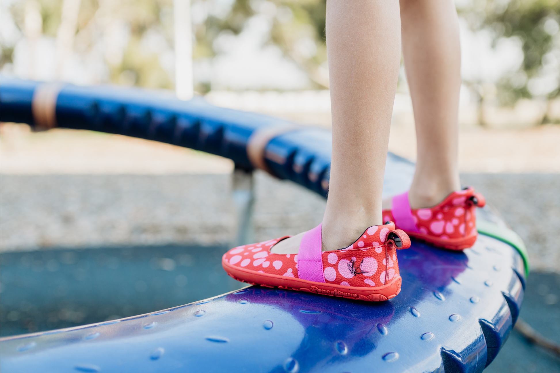 Child's feet wearing red leather PaperKrane barefoot shoes with pink dots on a blue playground structure.