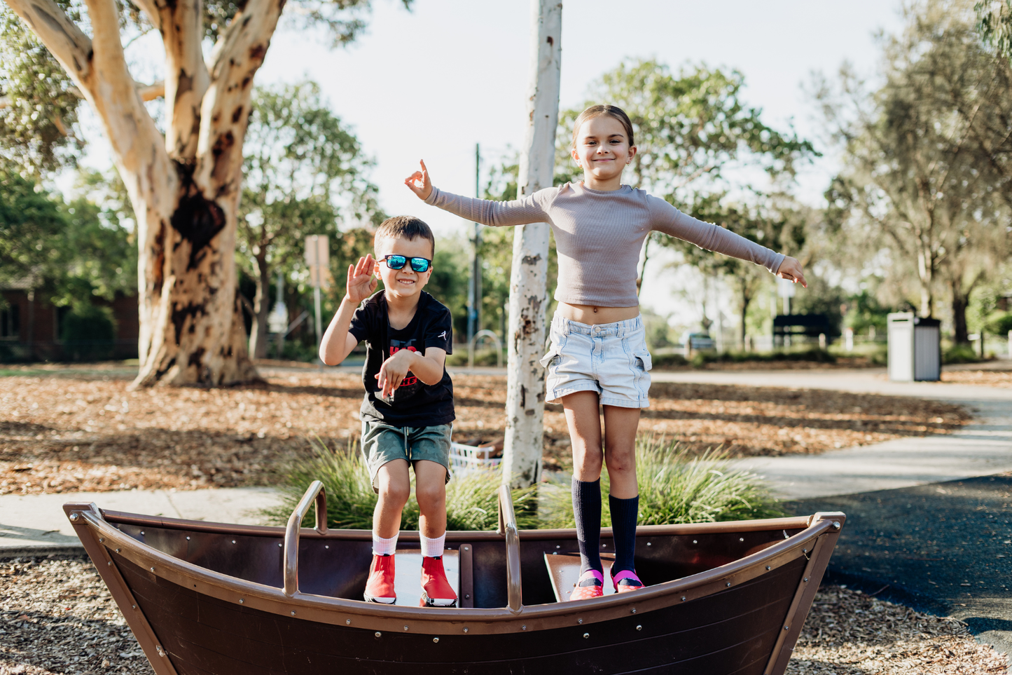 Two children jump and dance in a playground boat. The child on the left is wearing red leather PaperKrane barefoot chelsea boots with a zebra print rubber sole, and the child on the right is wearing red leather PaperKrane foot-shaped mary janes, with pink spots painted on them. 