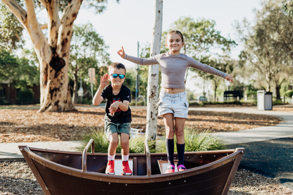 Two children jump and dance in a playground boat. The child on the left is wearing red leather PaperKrane barefoot chelsea boots with a zebra print rubber sole, and the child on the right is wearing red leather PaperKrane foot-shaped mary janes, with pink spots painted on them. 