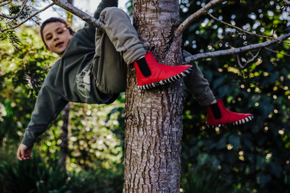 A child climbing a tree outdoors, wearing bright red leather foot-shaped chelsea boots with black and white striped soles, gray sweatpants, and a dark green hoodie.