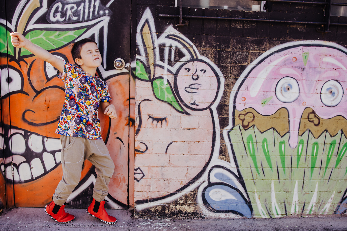 A young boy wearing a colorful patterned short-sleeve shirt, beige pants, and red leather PaperKrane barefoot chelsea boots with zebra soles, stands in front of a graffiti-covered wall, posing with one arm raised. 