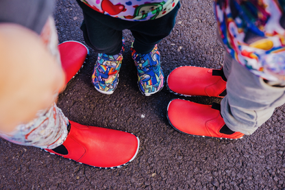 A top down view of a family in PaperKrane shoes, Mum and one child are in red leather chelsea boots with zebra soles, and the youngest is in the Graffiti print signature slip ons with zebra soles. A great way to match across our foot-shaped range of barefoot shoes!