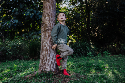 A young boy stands leaning against a tree trunk in a grassy, wooded outdoor setting. He is wearing a dark green long-sleeve shirt, beige cargo pants, and bright red leather PaperKrane chelsea boots.
