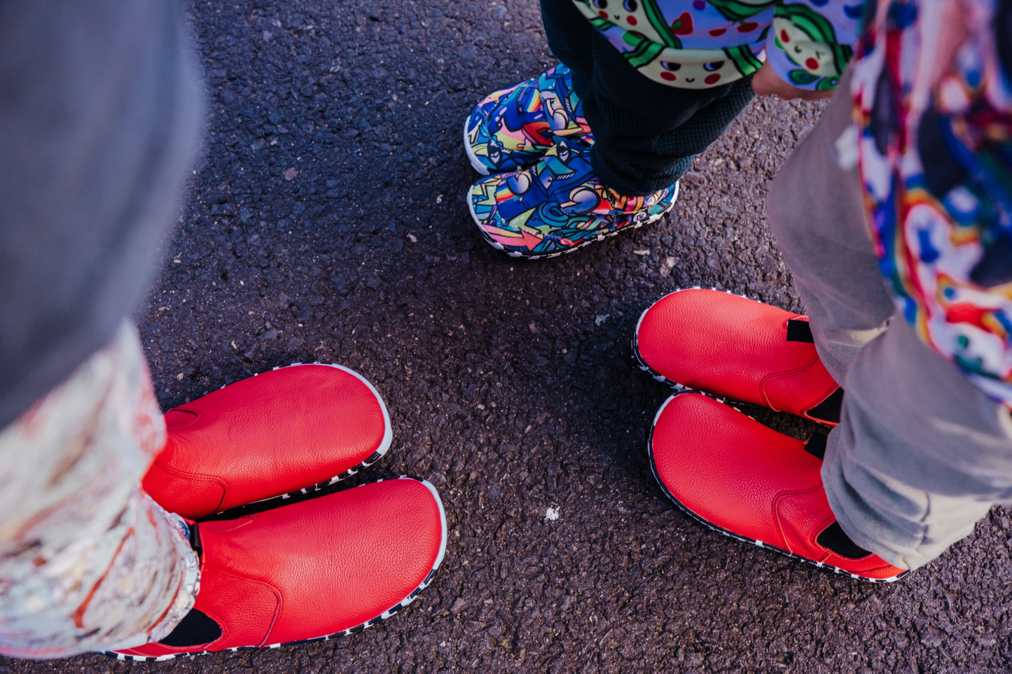 A top down view of a family in PaperKrane shoes, Mum and one child are in red leather chelsea boots with zebra soles, and the youngest is in the Graffiti print signature slip ons with zebra soles. They're standing on concrete showing the foot-shaped nature of PaperKrane boots and shoes. 