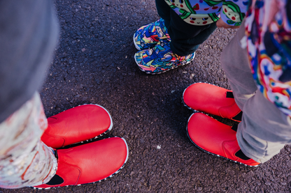 A top down view of a family in PaperKrane shoes, Mum and one child are in red leather chelsea boots with zebra soles, and the youngest is in the Graffiti print signature slip ons with zebra soles. They're standing on concrete showing the foot-shaped nature of PaperKrane boots and shoes. 