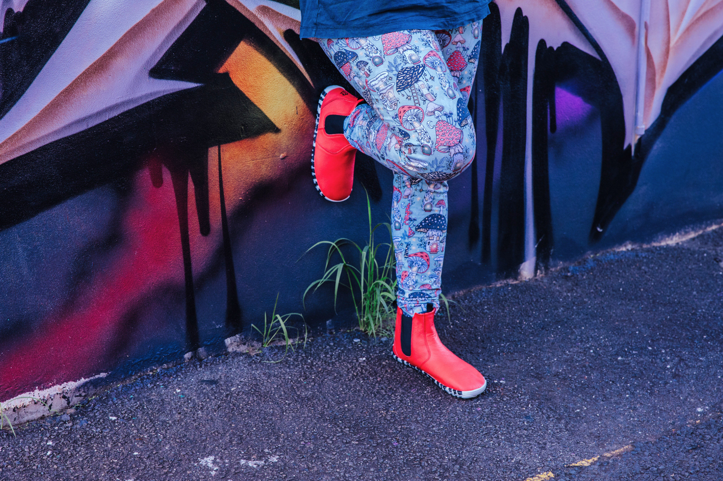 A woman in mushroom print leggings is leaning against a graffitied wall with one foot up. She's wearing red leather chelsea boots with zebra soles. One of PaperKrane's barefoot boots that are extra fun!