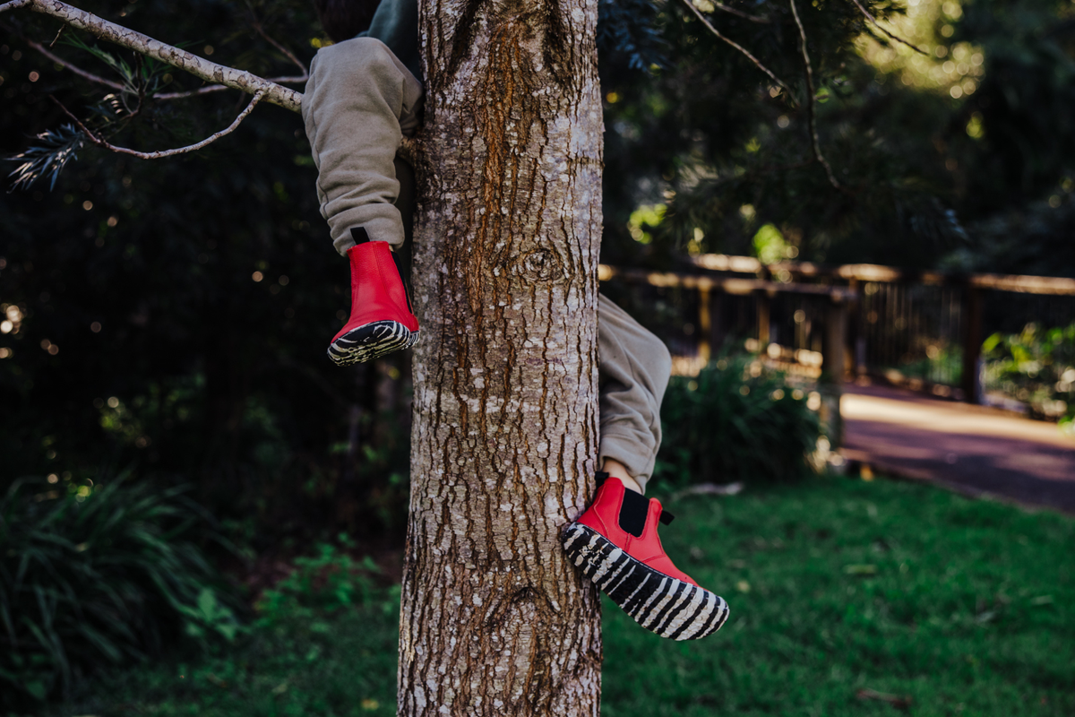 A child's legs in beige pants hang from a tree trunk outdoors, wearing bright red leather foot-shaped chelsea boots with black and white zebra-patterned soles.