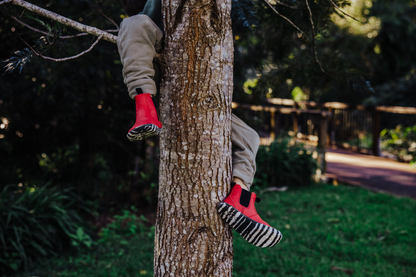 A child's legs in beige pants hang from a tree trunk outdoors, wearing bright red leather foot-shaped chelsea boots with black and white zebra-patterned soles.