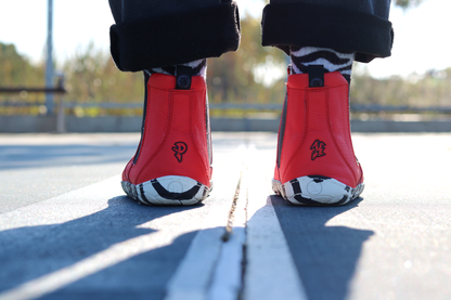 Red leather PaperKrane barefoot chelsea boots with a zebra print rubber sole. Viewed from the back on a person with zebra print socks standing on a court. 