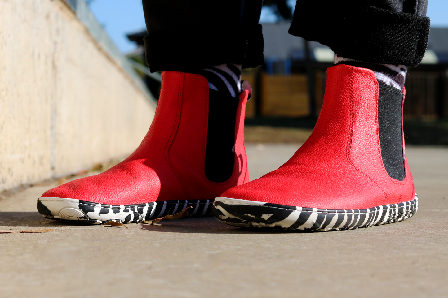 Red leather PaperKrane barefoot chelsea boots with a zebra print rubber sole. Viewed from a front angle, they have a foot-shaped toe box and are worn by a person with black pants and zebra socks, on concrete.