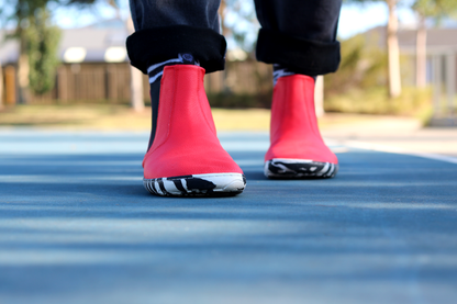 A person standing on a blue court wearing black pants and red leather PaperKrane barefoot chelsea boots with a zebra print rubber sole. 