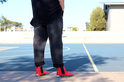 A person in black clothing standing next to a court, in red leather PaperKrane barefoot chelsea boots with a zebra print rubber sole. We can see the black embossed P and K logos on the back of the boots. 