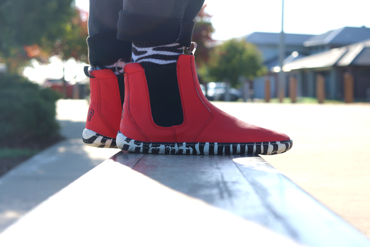Red leather PaperKrane barefoot chelsea boots with a zebra print rubber sole on a person with black pants and zebra socks. In an urban setting. 