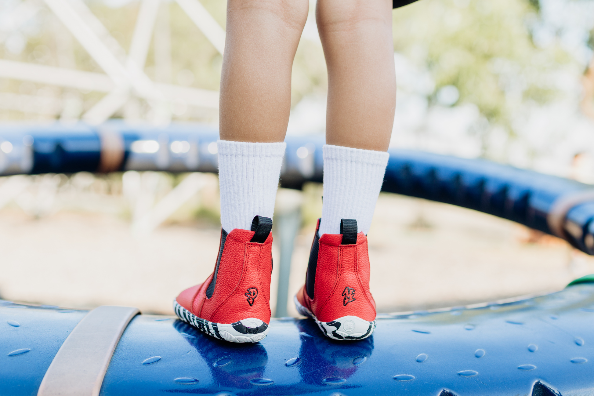 A child with long white socks is facing away from the camera to show the back of some red leather PaperKrane barefoot chelsea boots with a zebra print rubber sole. 