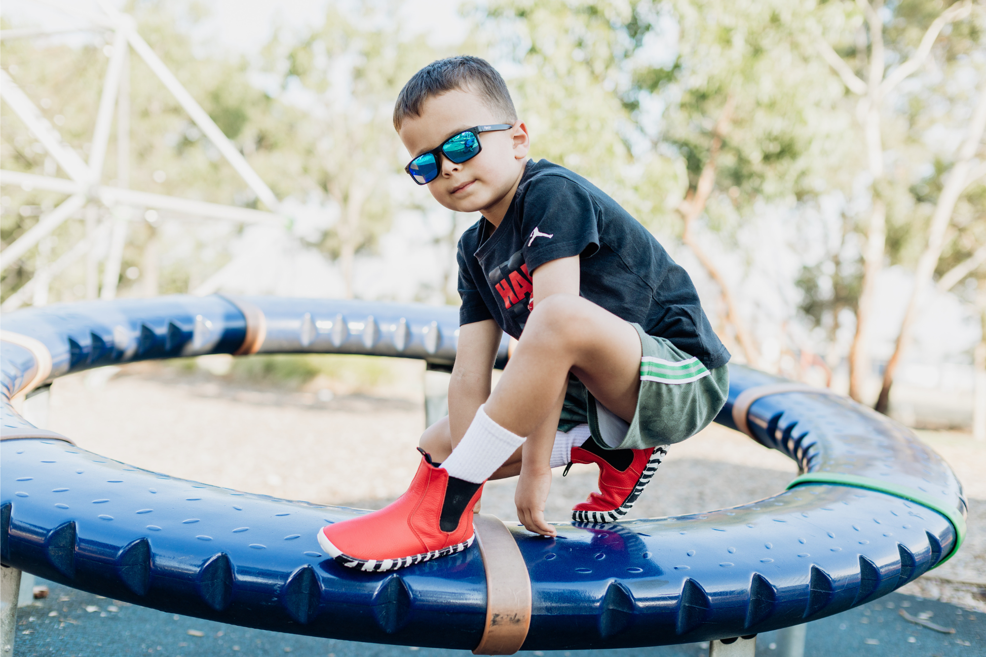 A child in sunglasses and tall white socks is crouched on a blue playground spinner, wearing red leather PaperKrane barefoot chelsea boots with a zebra print rubber sole. 