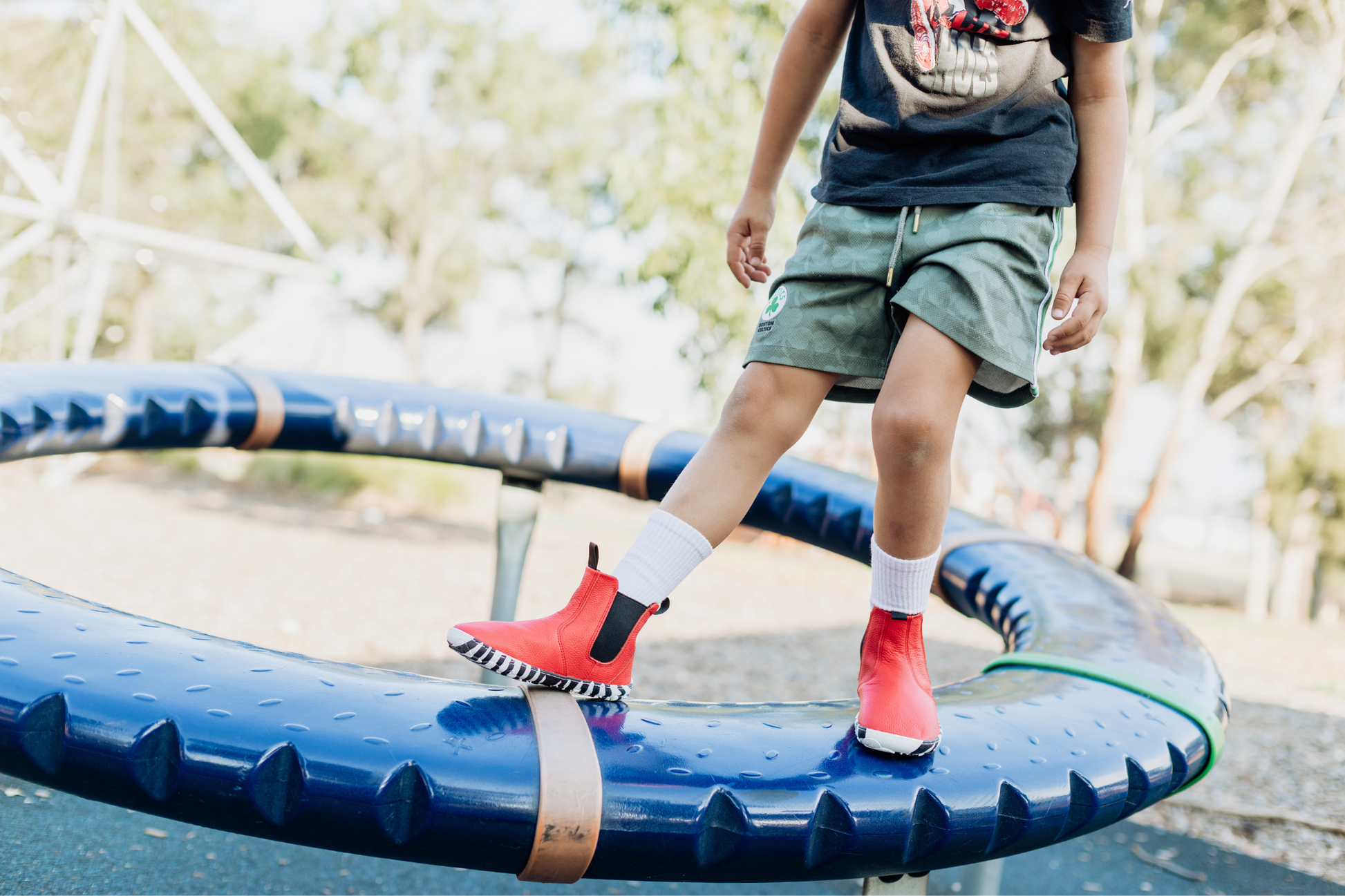 A child standing on a blue spinning playground thing is wearing long white socks in red leather PaperKrane barefoot chelsea boots with a zebra print rubber sole. 