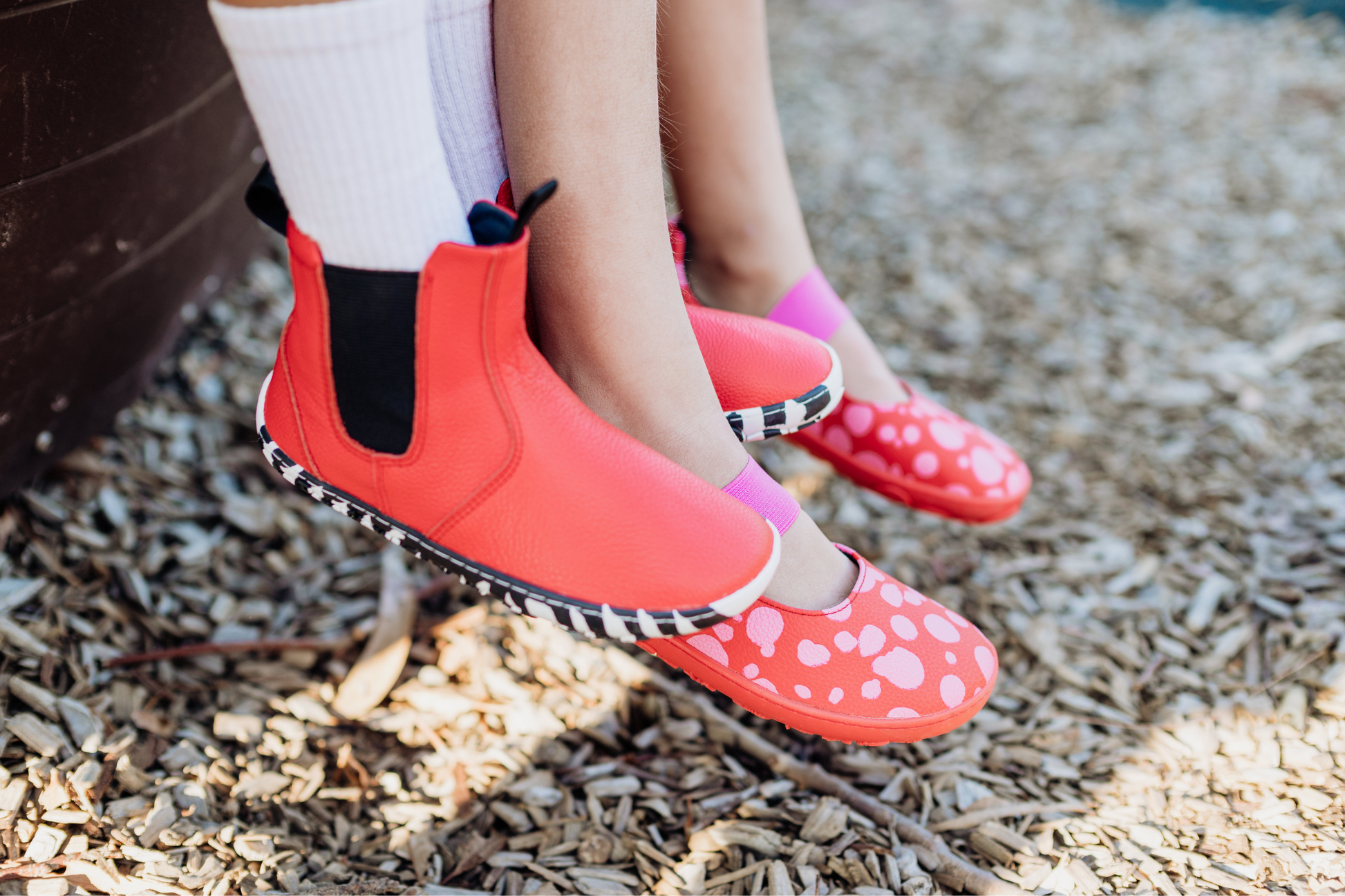 Red leather PaperKrane barefoot chelsea boots with a zebra print rubber sole and red leather foot-shaped mary janes from PaperKrane are worn on the feet of two children, the feet are intertwined. 
