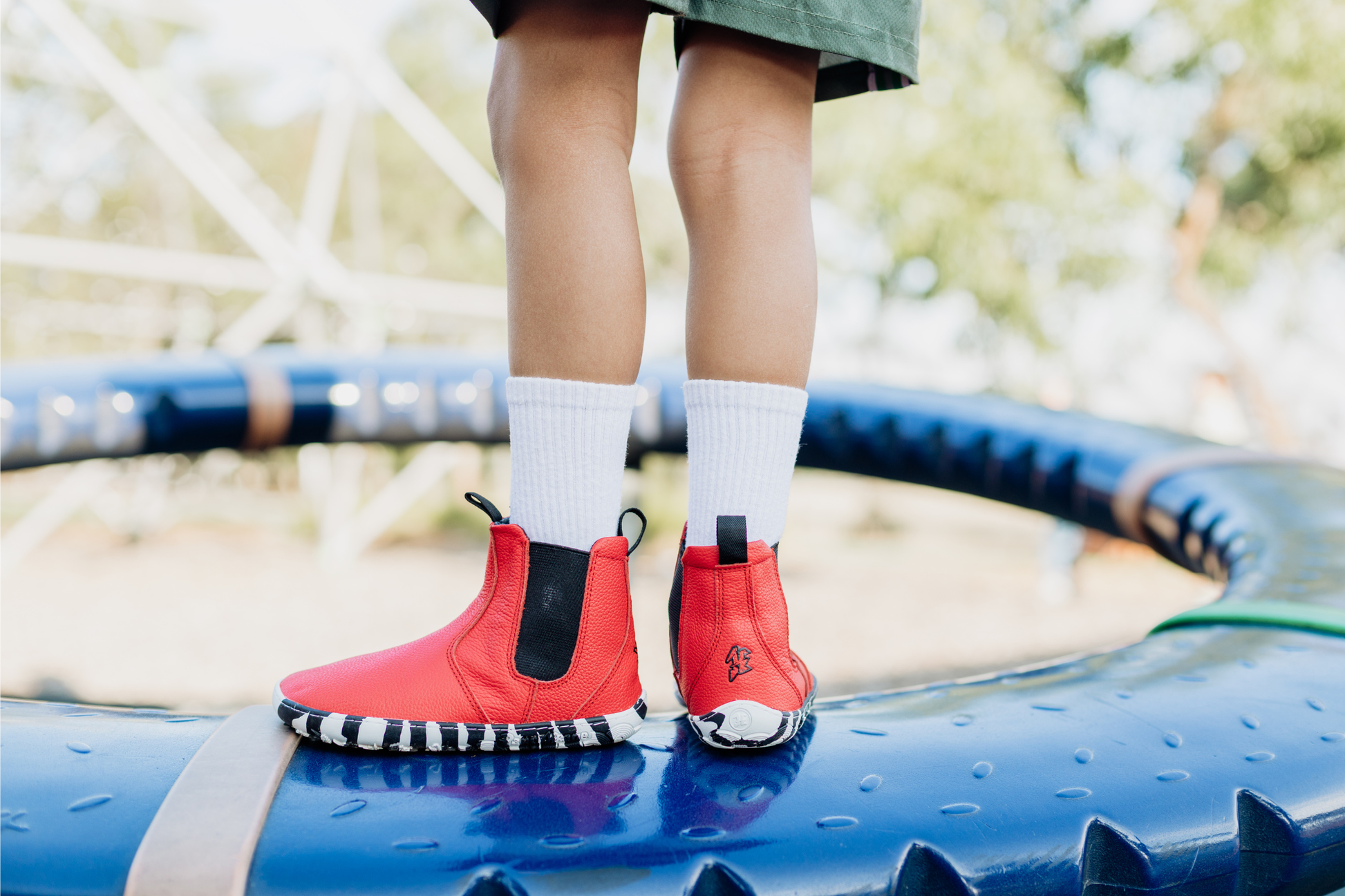 A child with white socks is wearing red leather PaperKrane barefoot chelsea boots with a zebra print rubber sole, they're standing on a piece of blue playground equipment with one boot turned to the left, and the back of the other boot facing the camera. 