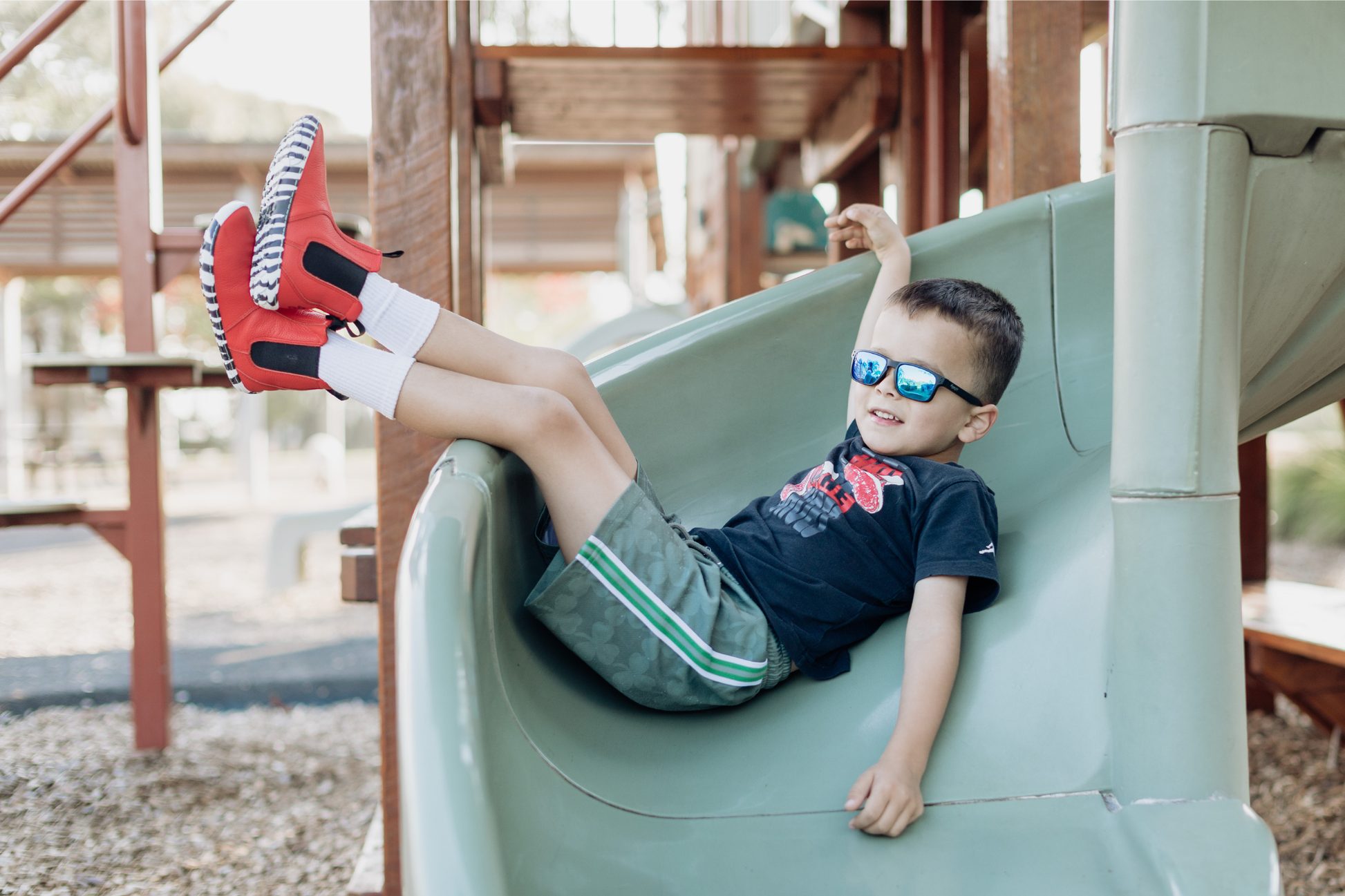 A child with sunglasses is lounging with his feet up, in a slide, wearing long white socks, and red leather PaperKrane barefoot chelsea boots with a zebra print rubber sole. 
