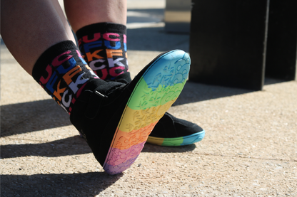 Person wearing colourful socks with letters and a rainbow-patterned shoe on a concrete surface.