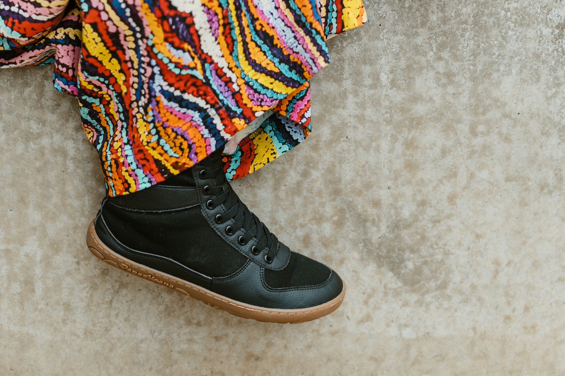 Adult leg in a barefoot PaperKrane SNUGG boot, the black vegan boots with elastic laces are paired with some colourful pants from the clothing brand OBUS, one leg dangles on a concrete wall. 