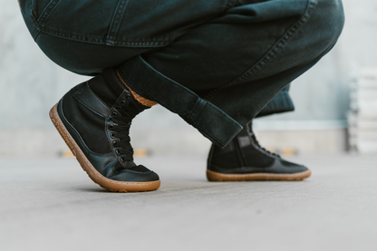 Adult in black work pants is crouching in a pair of black vegan foot-shaped PaperKrane SNUGG boots, on concrete. 